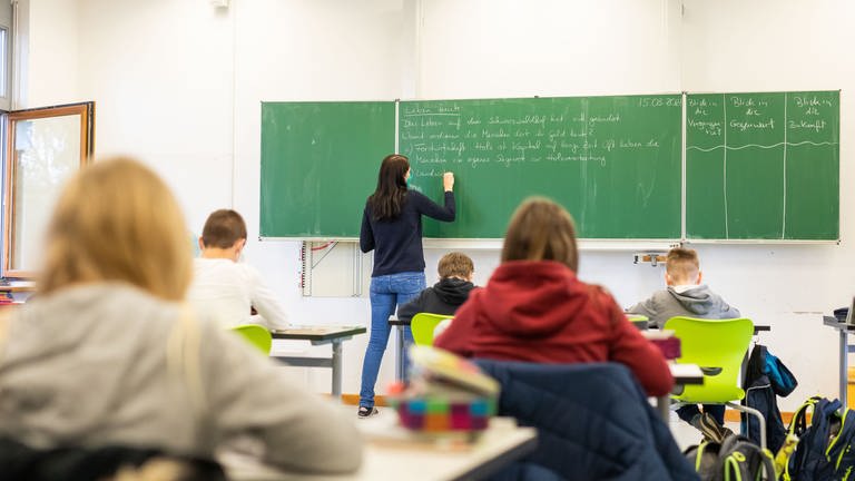 Eine Lehrerin steht an einer Tafel in einem Klassenzimmer, Schüler lernen. Ab dem kommenden Wintersemester können Studierende in einem Modellversuch in einem dualen Masterstudiengang auf Lehramt studieren. 
